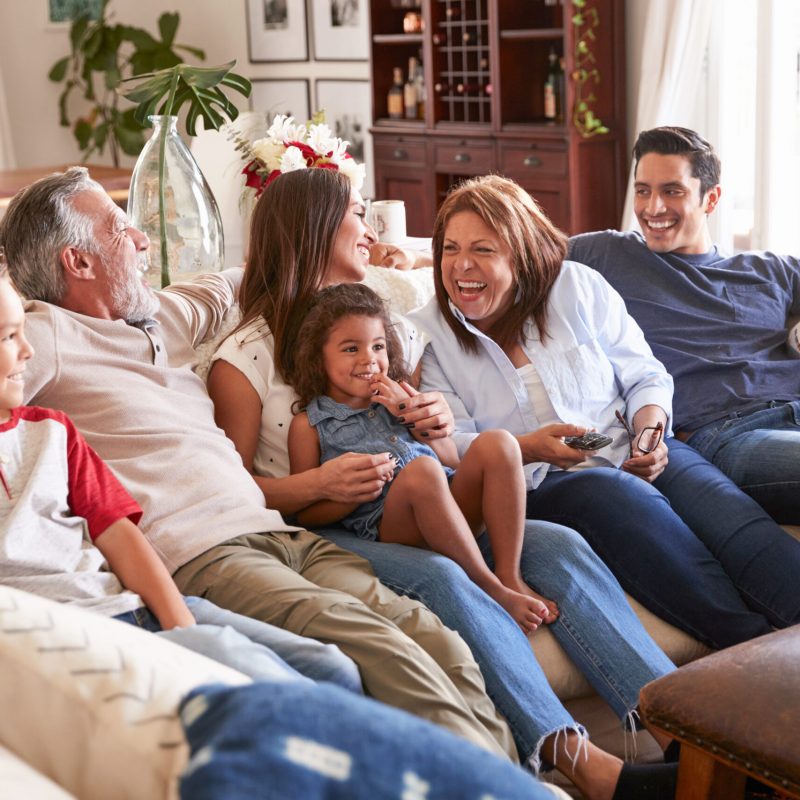 Three generation Hispanic family sitting on the sofa watching TV, grandmother using remote control; Shutterstock ID 1285072144; purchase_order: Customer contribution; job: 4706510; client: Sonova Australia ; other: Sonova